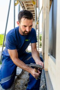 Handwerker mit Hammer an Holzkonstruktion der Hausfassade: Ein Lüftungsprofil als Sockelabschluss erhält den Kamineffekt der Hinterlüftung und schützt den Zwischenraum vor Ungeziefer.