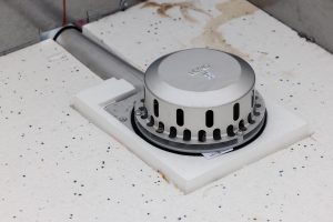 Der Wärmedämmblock ist auf die Form des Abflusses angepasst. Es muss nur ein quaderförmiger Block aus dem EPS geschnitten werden – und der Abfluss ist formschlüssig in der Dämmebene. Das spart Arbeitszeit. Die Hauptentwässerung führt dann durch die Attika und in der Luftschicht der VHF nach unten.