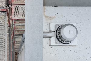 Der Wärmedämmblock ist auf die Form des Abflusses angepasst. Es muss nur ein quaderförmiger Block aus dem EPS geschnitten werden – und der Abfluss ist formschlüssig in der Dämmebene. Das spart Arbeitszeit. Die Hauptentwässerung führt dann durch die Attika und in der Luftschicht der VHF nach unten.