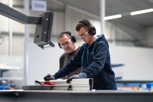 In der Werkshalle von TS Aluminium: Auszubildender und Begleiter beim Zuschneiden von Aluminiumprofilen an der Schneidemaschine. - Der Workshop „Dachzuschnitt“ findet direkt „an der Säge“ in der Fertigung statt.