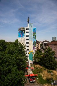 Außenaufnahme von Malerarbeiten an der Hochhaus-Fassade mit Hebebühne. - Das Mural „In Contemplation“ von vom Künstler TAXIS in seiner Entstehung an der Fassade eines Wohn-Hochhauses im Mannheimer Stadtteil Lindenhof. Zwei Wochen lang verwandelten er und sein Team die Wände mit bunten Fassadenfarben, Rollen und Pinseln in eine farbenfrohe Landschaft.
