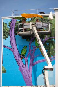 Auf Hebebühne arbeiten Künstler unter Sonnenschutz an dem Kunstwerk an einer Hochhaus-Fassade. – Das Mural „In Contemplation“ von vom Künstler TAXIS in seiner Entstehung an der Fassade eines Wohn-Hochhauses im Mannheimer Stadtteil Lindenhof. Zwei Wochen lang verwandelten er und sein Team die Wände mit bunten Fassadenfarben, Rollen und Pinseln in eine farbenfrohe Landschaft.